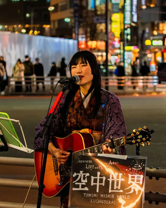 a busker in a purple kimono sings love and peace under the neon