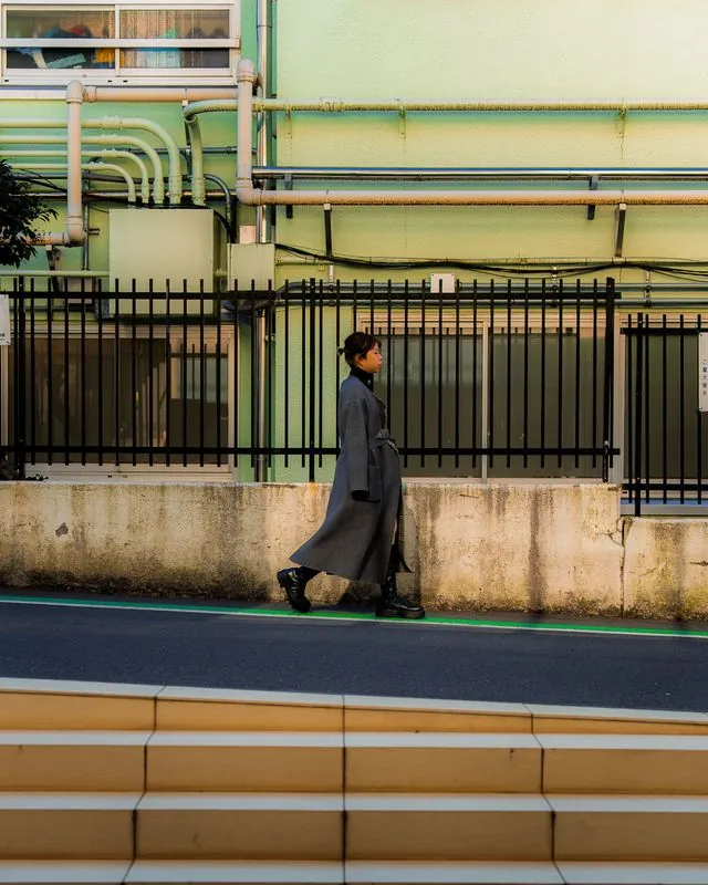 a woman in a gray coat walks past the pipes, mid-stride