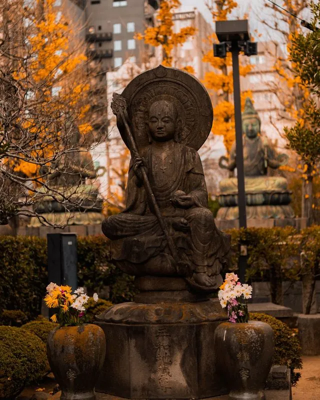golden autumn leaves frame the stone jizo, apartments looming behind