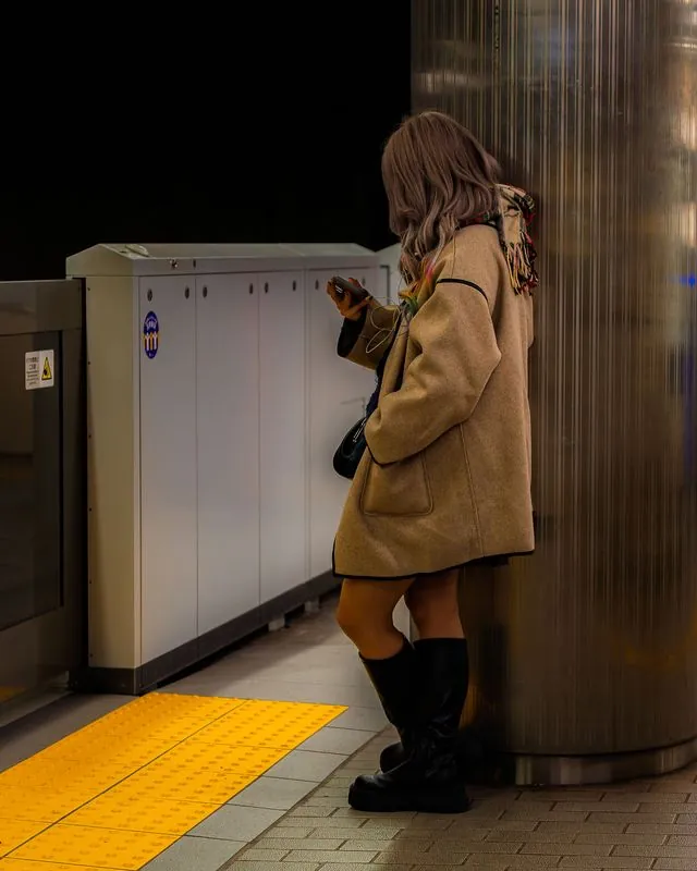 leaning on the steel column, she scrolls; the platform edge glows yellow behind her